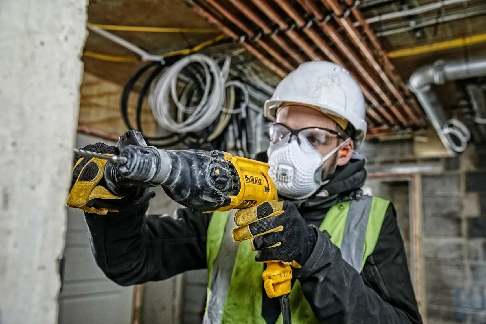 Un trabajador de la construcción con casco, gafas de seguridad, guantes y chaleco reflectante utiliza un martillo rotativo DEWALT D25033 en una obra. Al fondo se observan paredes sin terminar, tuberías y cables eléctricos.