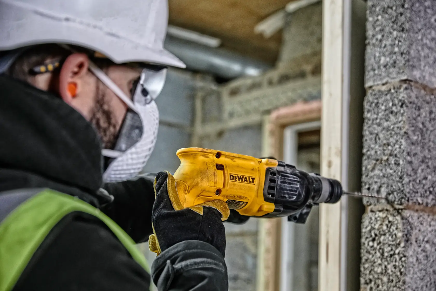Una persona con equipo de seguridad utiliza un taladro amarillo DEWALT para perforar una pared de hormigón en un sitio de construcción.