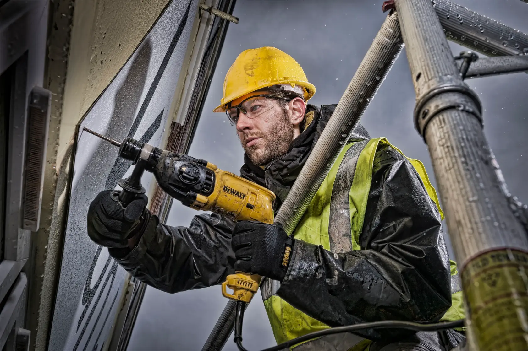 Un trabajador de construcción con casco amarillo y chaleco de seguridad utiliza un taladro percutor DEWALT (modelo D25033) para perforar una pared desde un andamio. La cara del trabajador está desenfocada para preservar su privacidad.