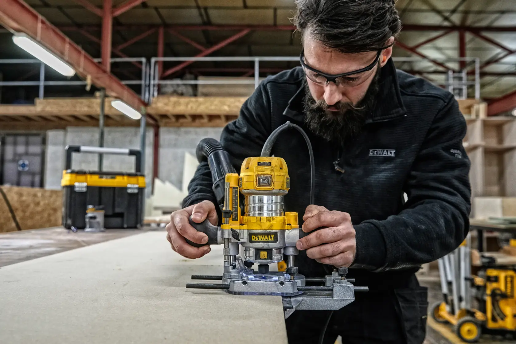Eine Person verwendet ein gelbes DEWALT D26204K Fräswerkzeug auf einem Holzstück in einer Werkstatt. Das Gesicht der Person ist unkenntlich gemacht, und im Hintergrund sind verschiedene Werkstatteinrichtungen und Aufbewahrungsboxen zu sehen.