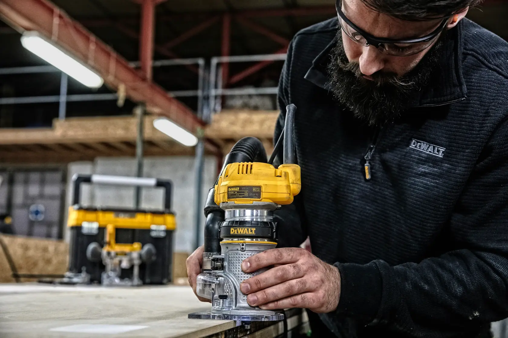 Eine Person verwendet ein DEWALT D26204K Fräswerkzeug auf einer Holzoberfläche in einer Werkstatt, wobei das Gesicht der Person aus Datenschutzgründen verschwommen ist. Im Hintergrund ist ein weiteres DEWALT Werkzeug zu sehen.
