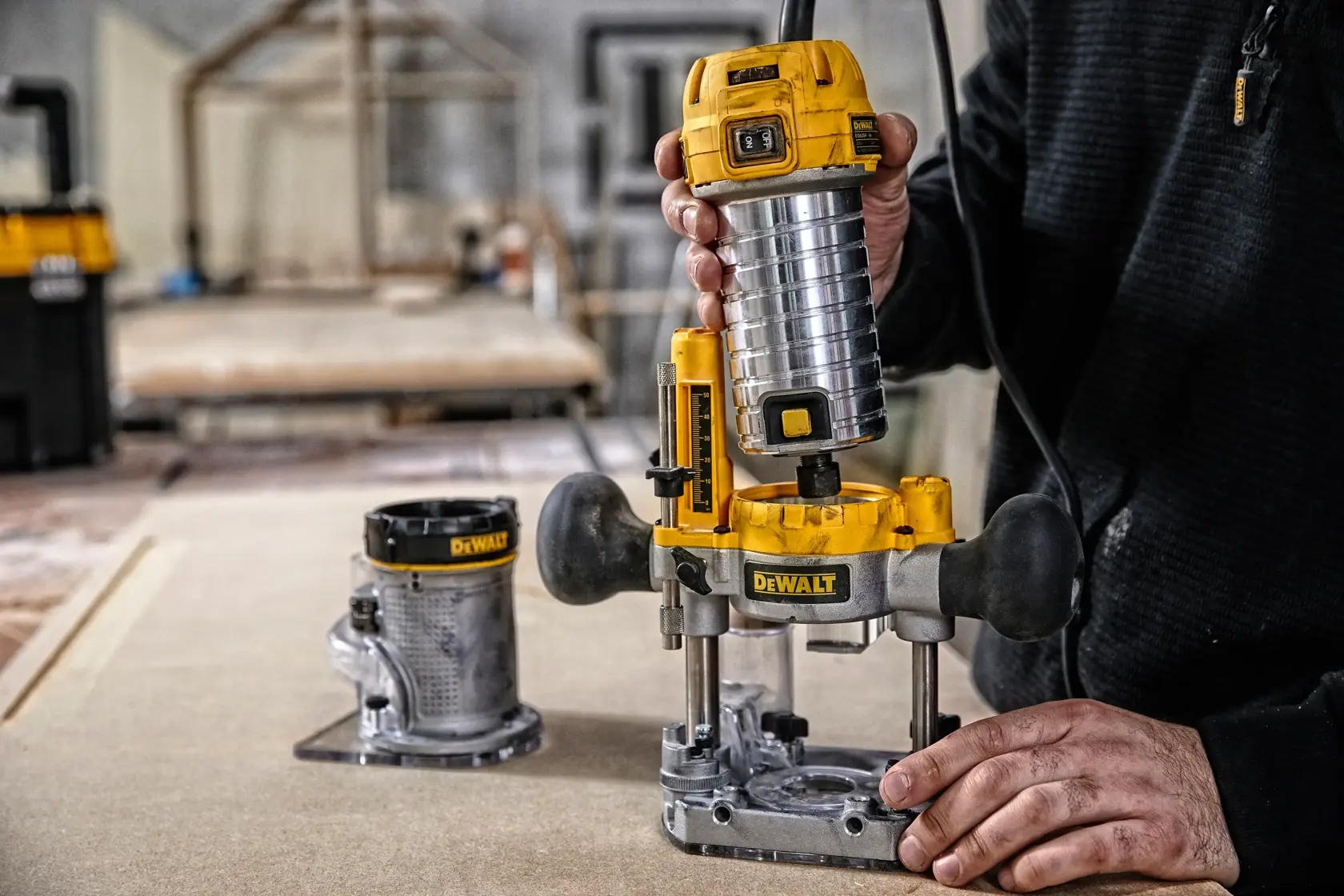 Eine Person verwendet ein DEWALT D26204K Fräswerkzeug auf einem Holzstück in einer Werkstatt. Eine weitere Basisbefestigung ist auf dem Tisch sichtbar. Der Fokus liegt auf dem gelb-silbernen DEWALT Fräswerkzeug, das bedient wird.