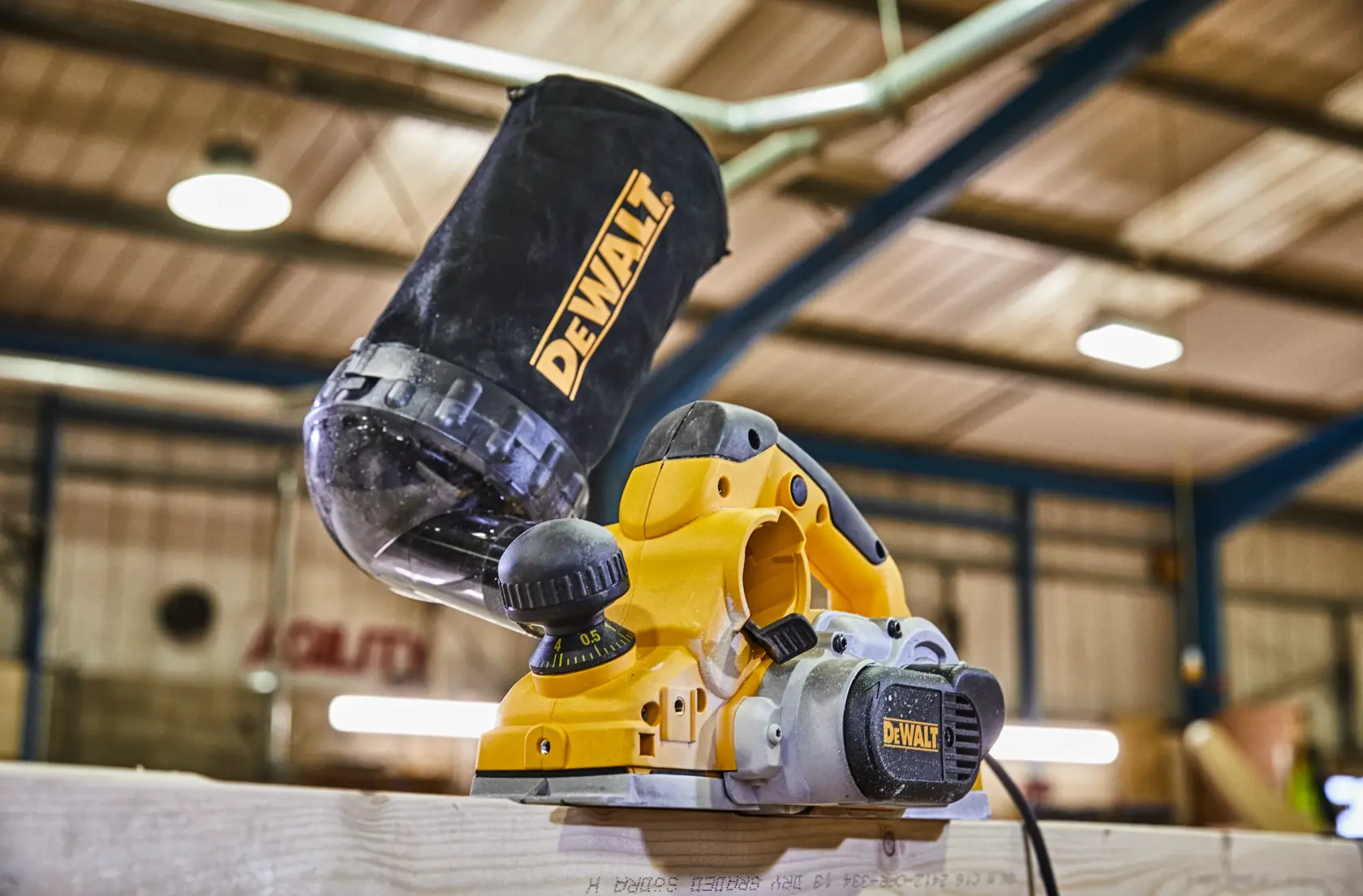 Ein elektrischer Hobel von DEWALT mit angeschlossenem Staubauffangbeutel, im Einsatz auf einem Holzbrett in einer Werkstatt.