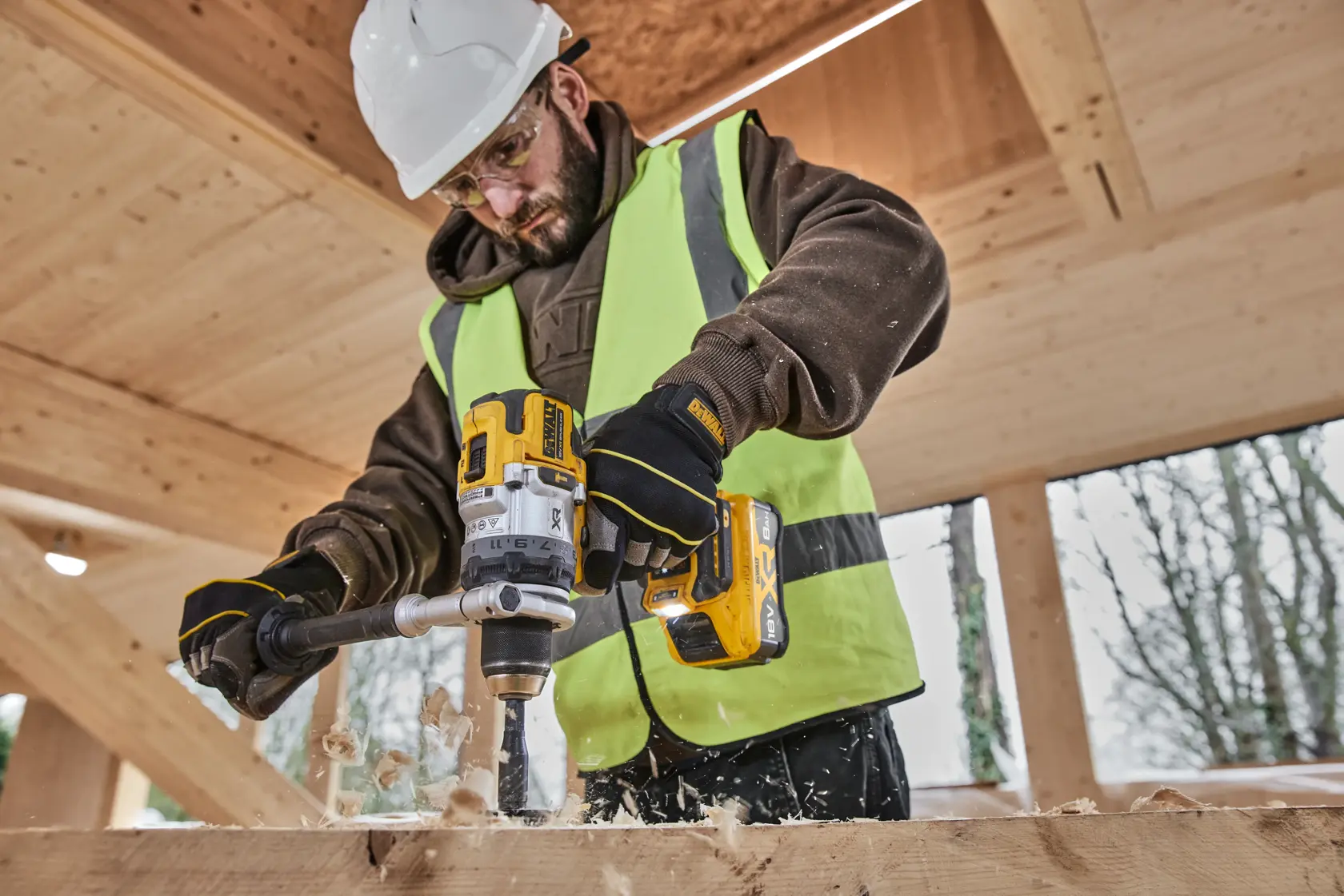 Persona perfora madera con DEWALT DCB1880. Lleva chaleco, guantes y casco en obra.