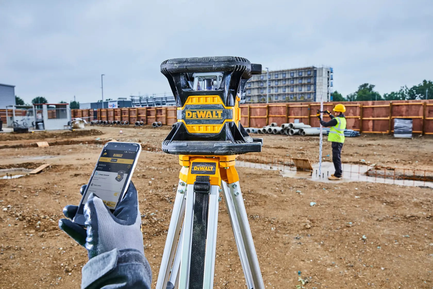 Ein DEWALT-Laser-Nivelliergerät auf einem Stativ auf einer Baustelle, mit einem Arbeiter im Hintergrund und einer Person mit einem Smartphone im Vordergrund.