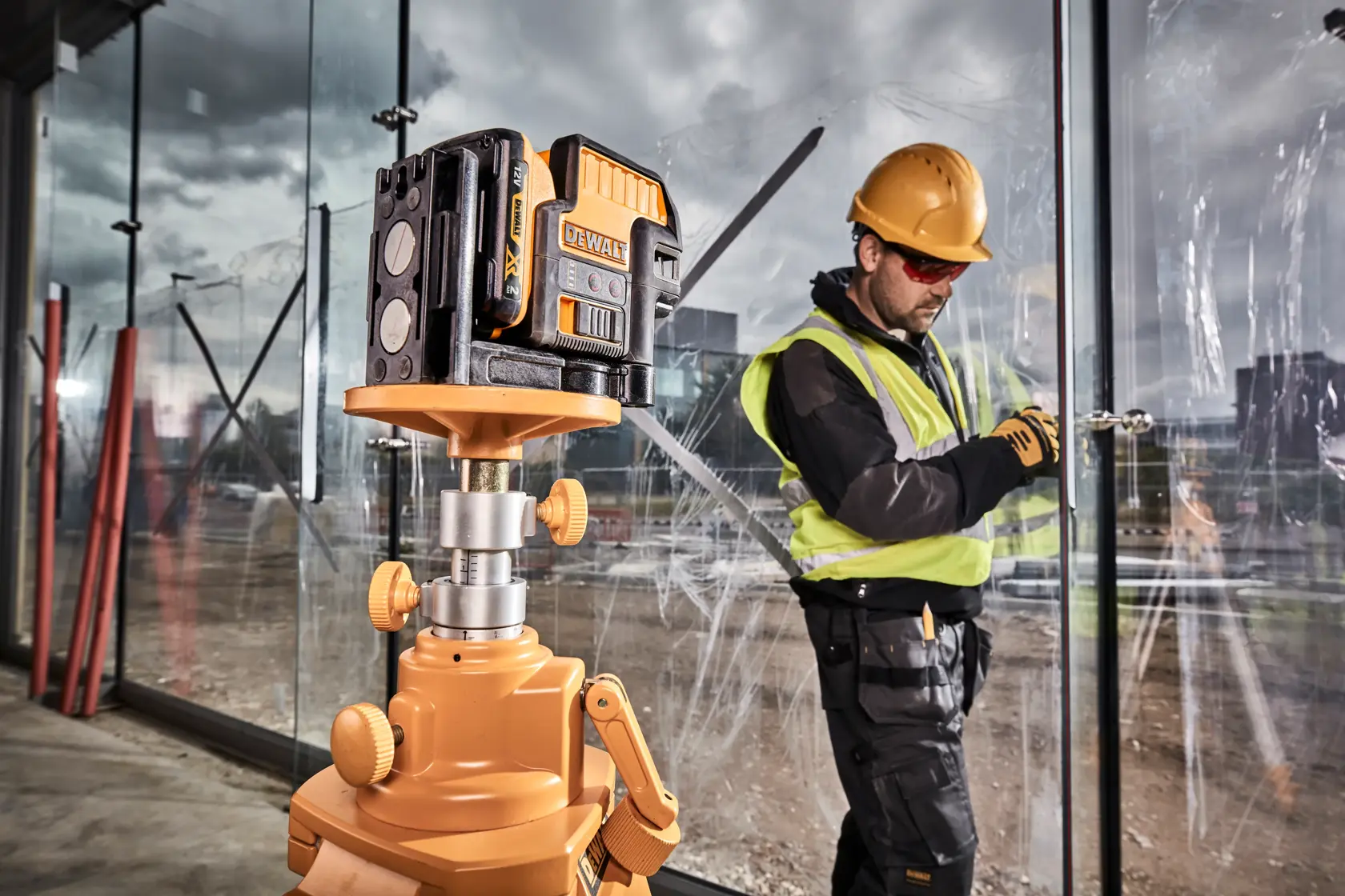 Ein DEWALT-Laser-Nivelliergerät ist auf einem Stativ auf einer Baustelle montiert, im Hintergrund arbeitet eine Person mit Sicherheitsweste und Schutzhelm in der Nähe großer Glasscheiben.