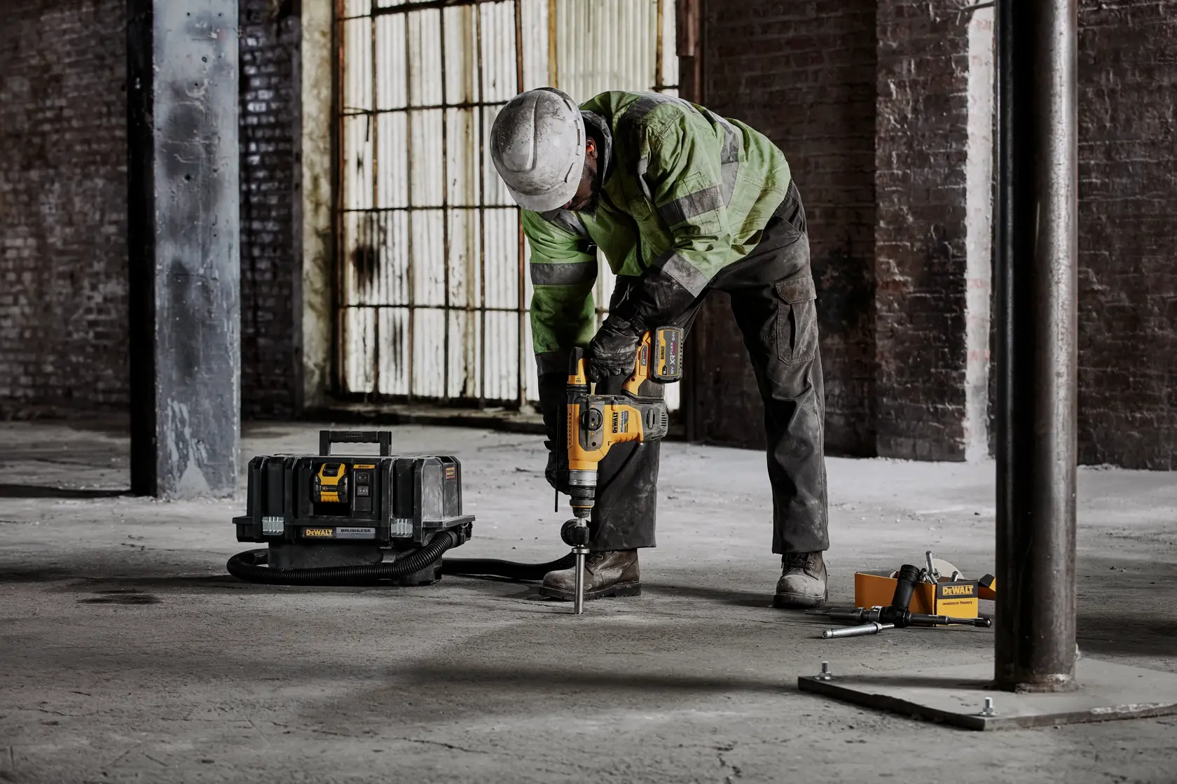 Persona usa martillo rotativo DEWALT en suelo de concreto, caja de herramientas cerca en obra.