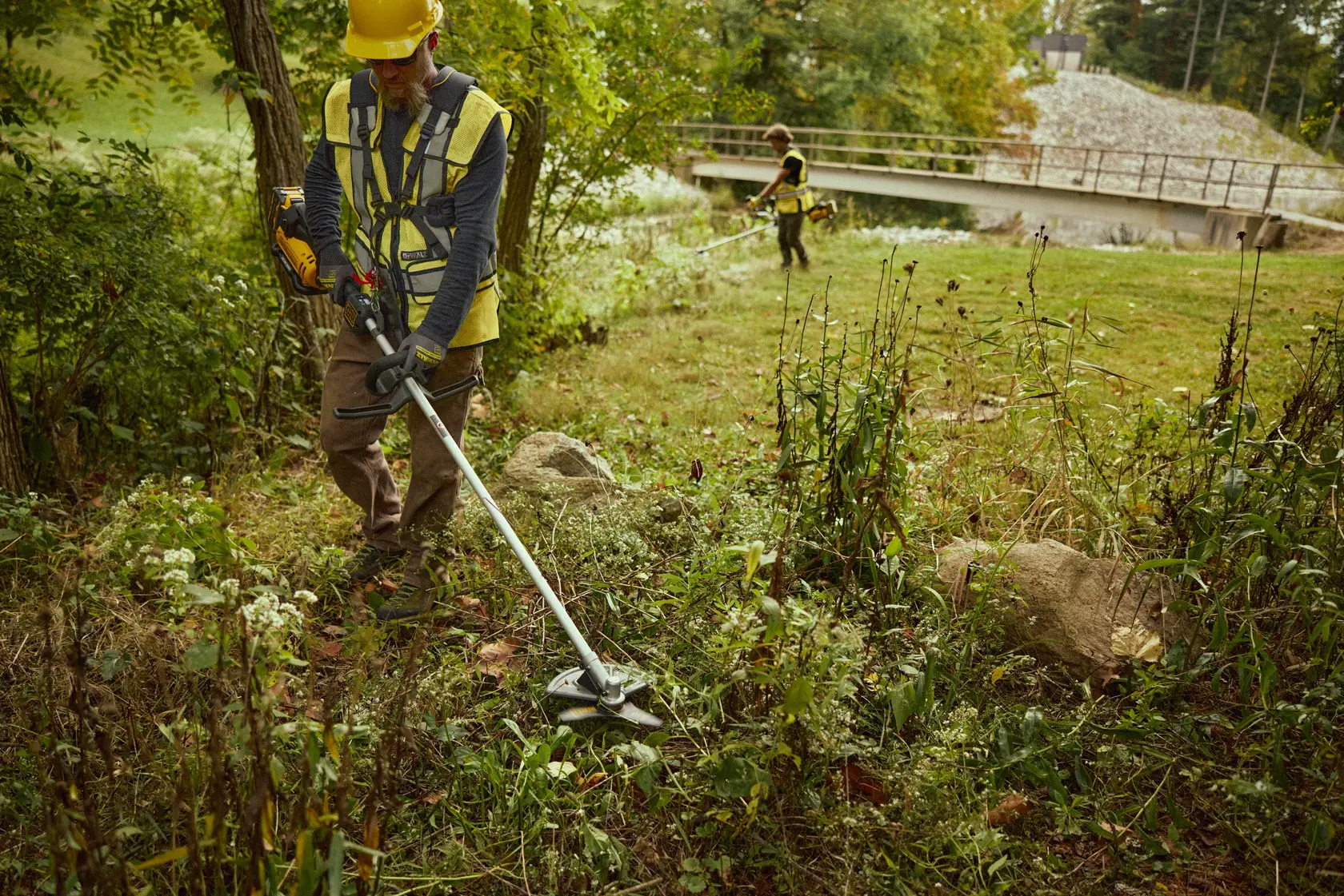 Zwei Personen trimmen Gras mit DEWALT DCMBC812N-XJ und DCMBC812N Freischneidern im Freien bei Bäumen sowie Brücke.
