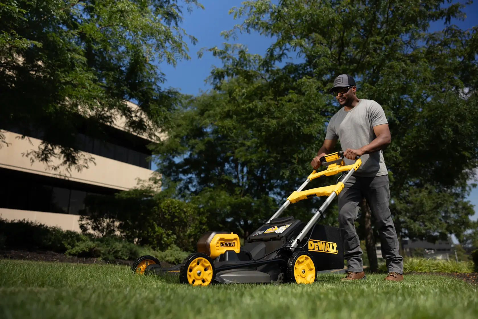 Une personne utilise une tondeuse DEWALT sur l’herbe, avec des bâtiments et arbres en arrière-plan.