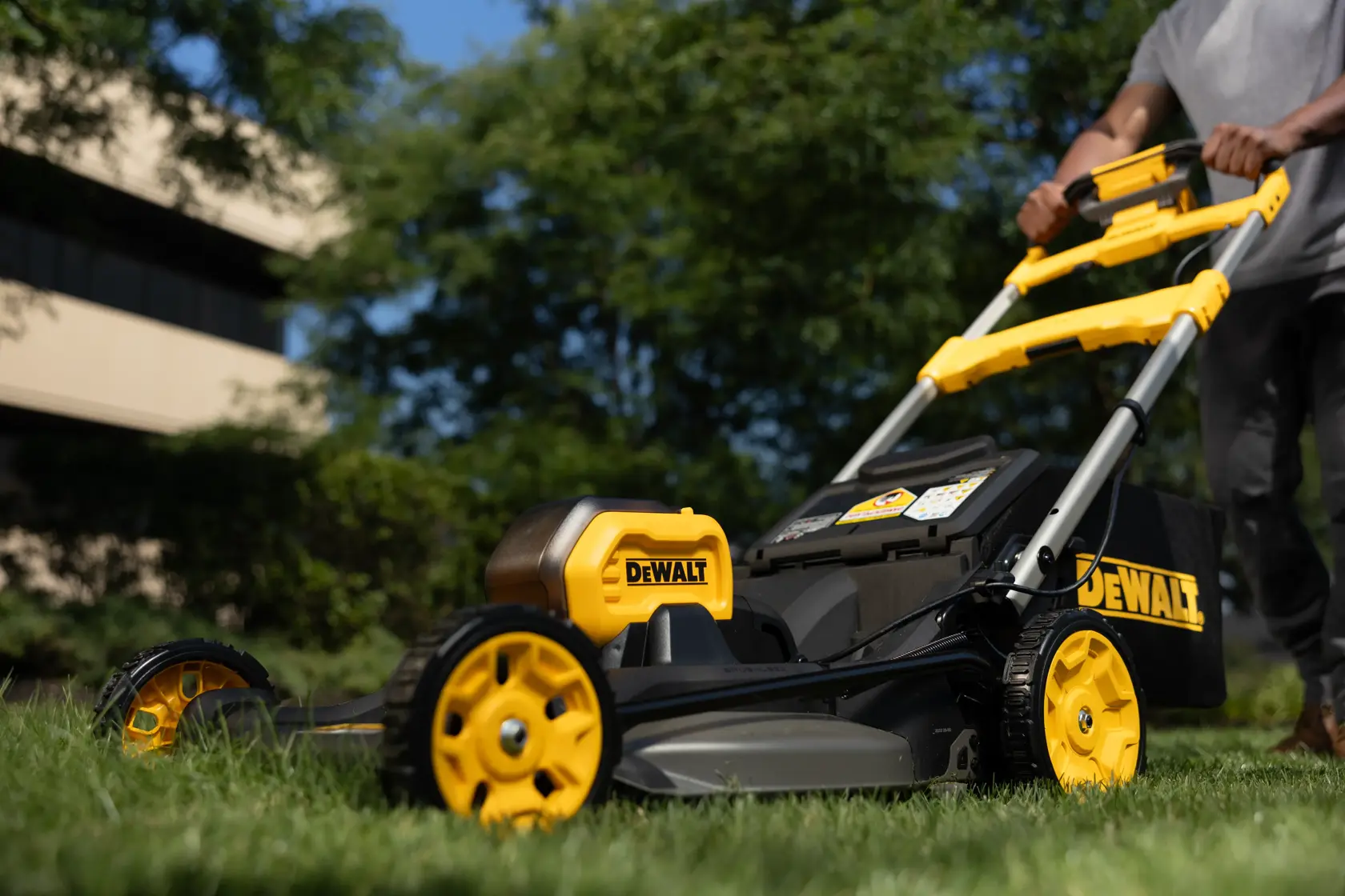 Une tondeuse à gazon DEWALT est poussée sur l’herbe, avec arbres et bâtiment en arrière-plan.