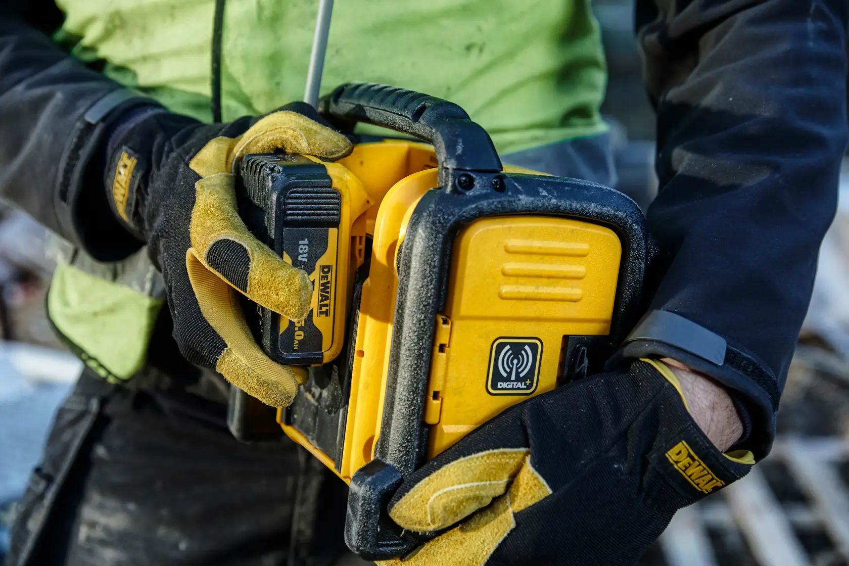 Person hält gelbes DEWALT DCR020 Digitalradio mit 18V Akku, trägt Arbeitshandschuhe auf Baustelle.