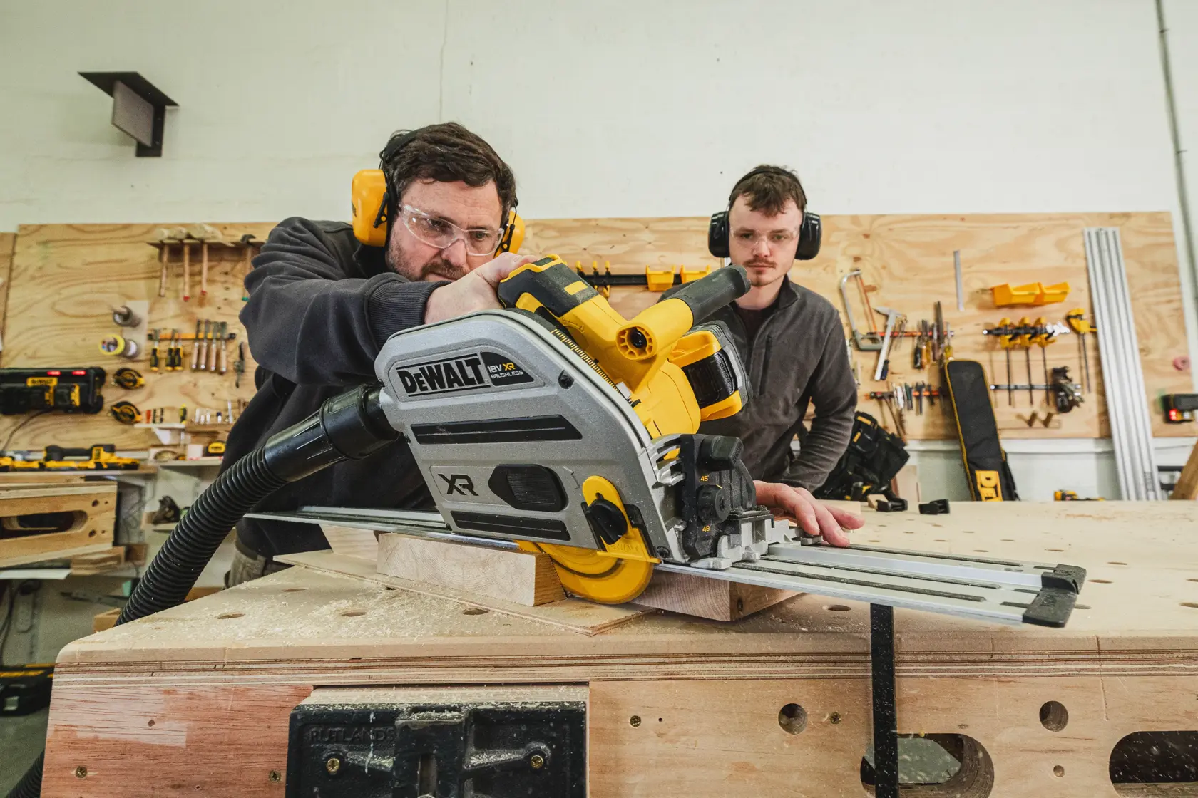 Zwei Personen schneiden Holz mit einer DEWALT XR Akku-Säge auf einer Werkbank in einer Werkstatt.