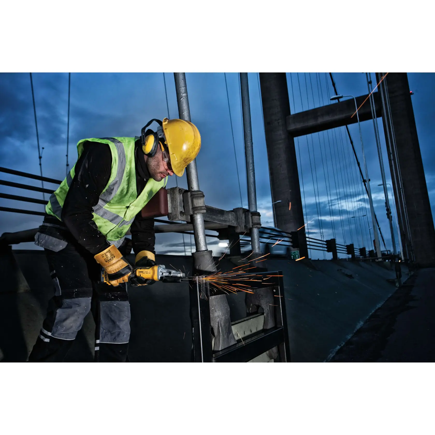 Trabajador con equipo de seguridad usa herramienta DEWALT en metal, chispas junto a un puente al atardecer.