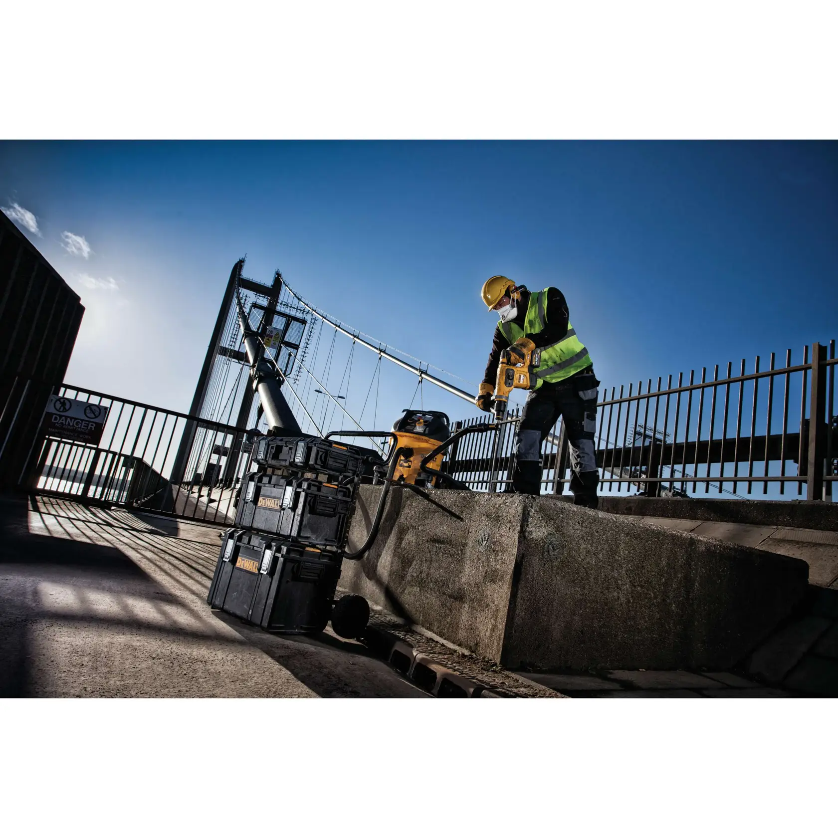 Trabajador usa herramienta DEWALT junto a cajas apiladas en obra exterior con puente de fondo.