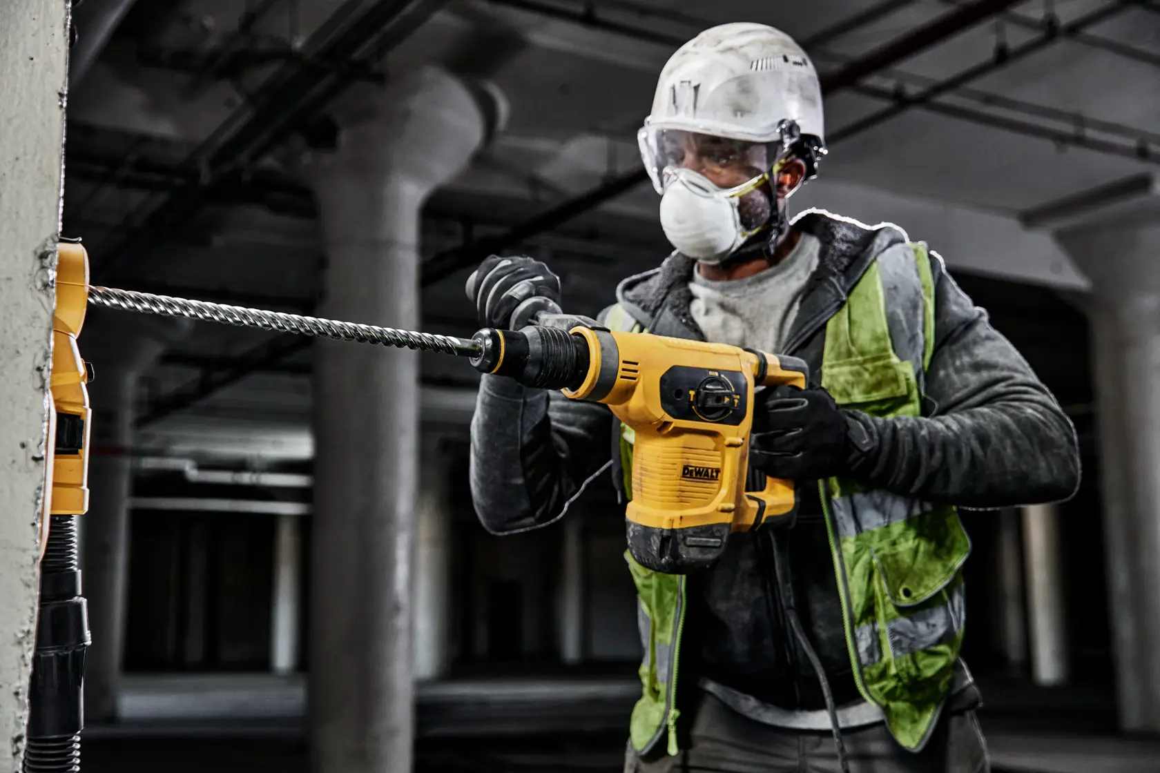 Trabajador usa taladro DEWALT en muro de concreto, viste casco y chaleco de seguridad.