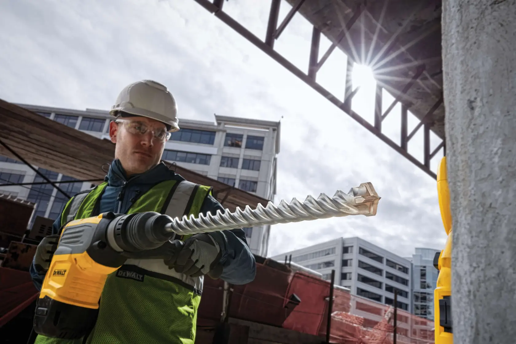 Trabajador usando taladro DEWALT con broca grande en obra, lleva protección, edificios de fondo.