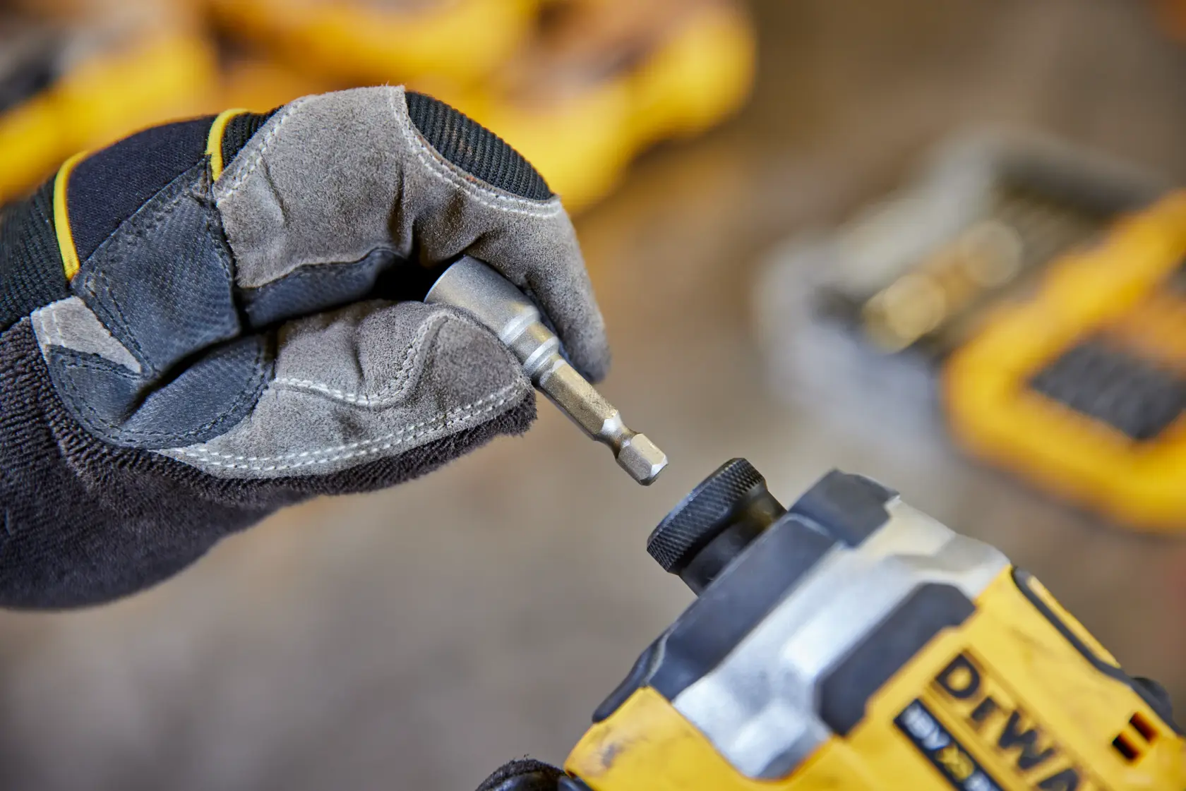 Mano enguantada coloca una punta DEWALT en herramienta amarilla. Hay kits borrosos al fondo.