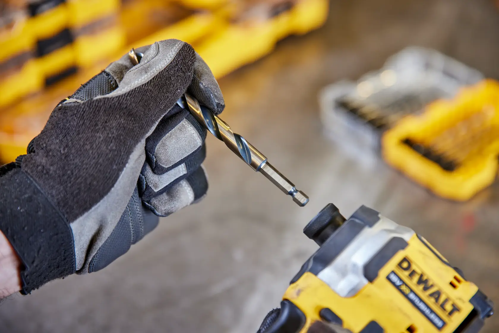 Mano con guante sostiene broca cerca de herramienta DEWALT con caja amarilla al fondo.