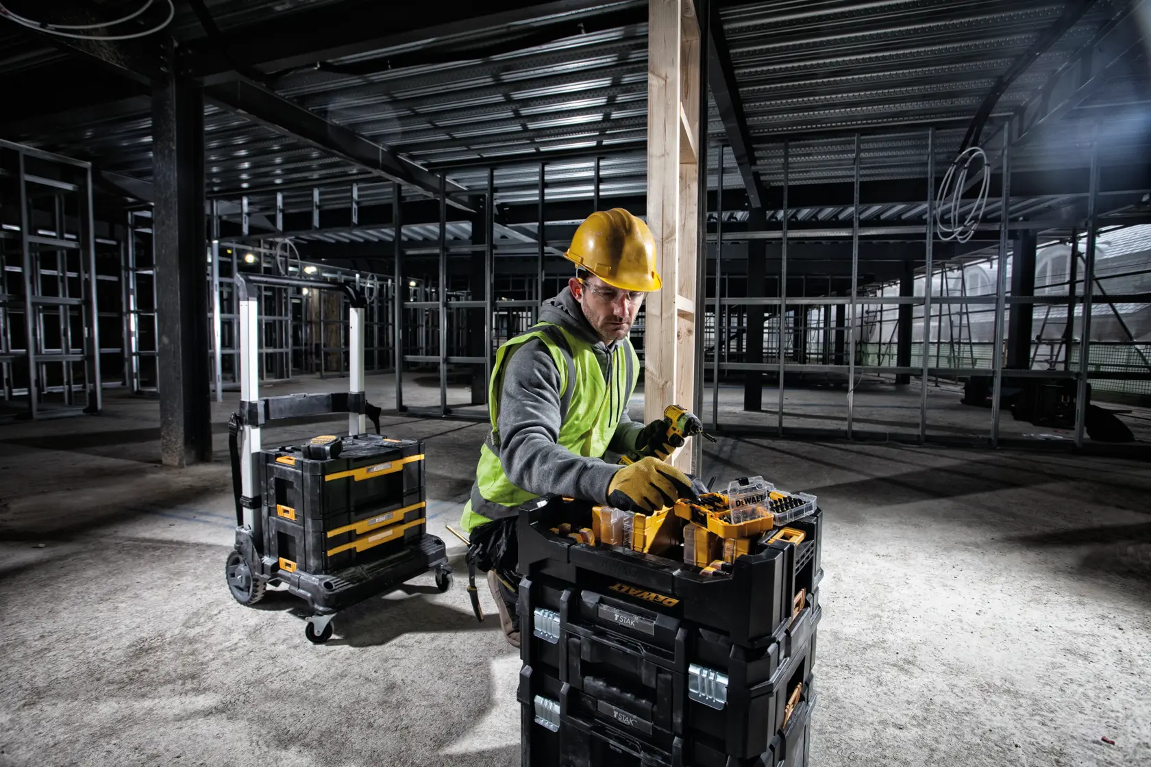 Trabajador con equipo de seguridad usando cajas DEWALT en una obra con estructuras metálicas y paredes sin terminar.