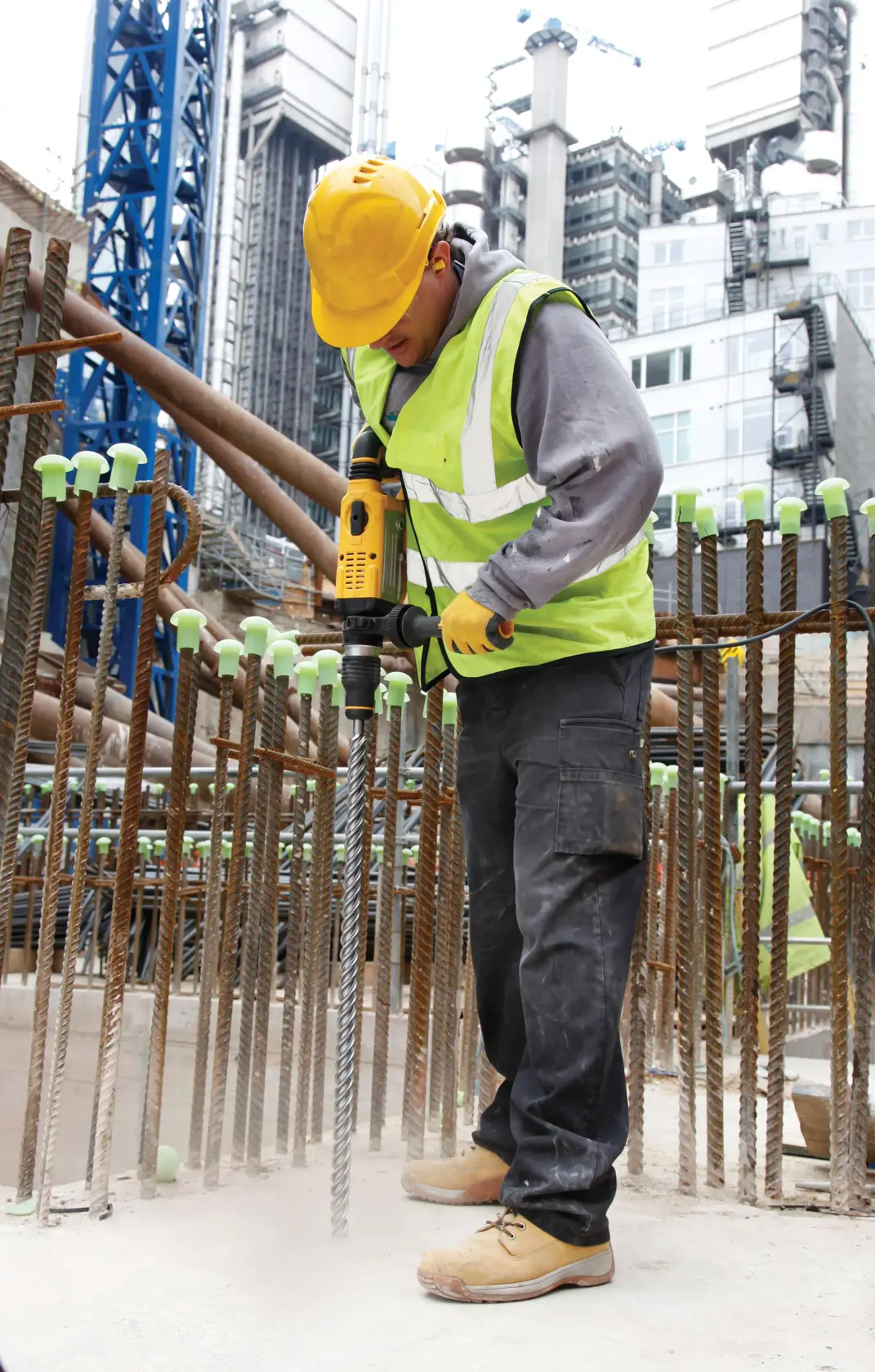 Arbeiter mit DEWALT-Bohrmaschine auf Baustelle, trägt Schutzkleidung, umgeben von Baustahl und Gebäuden.