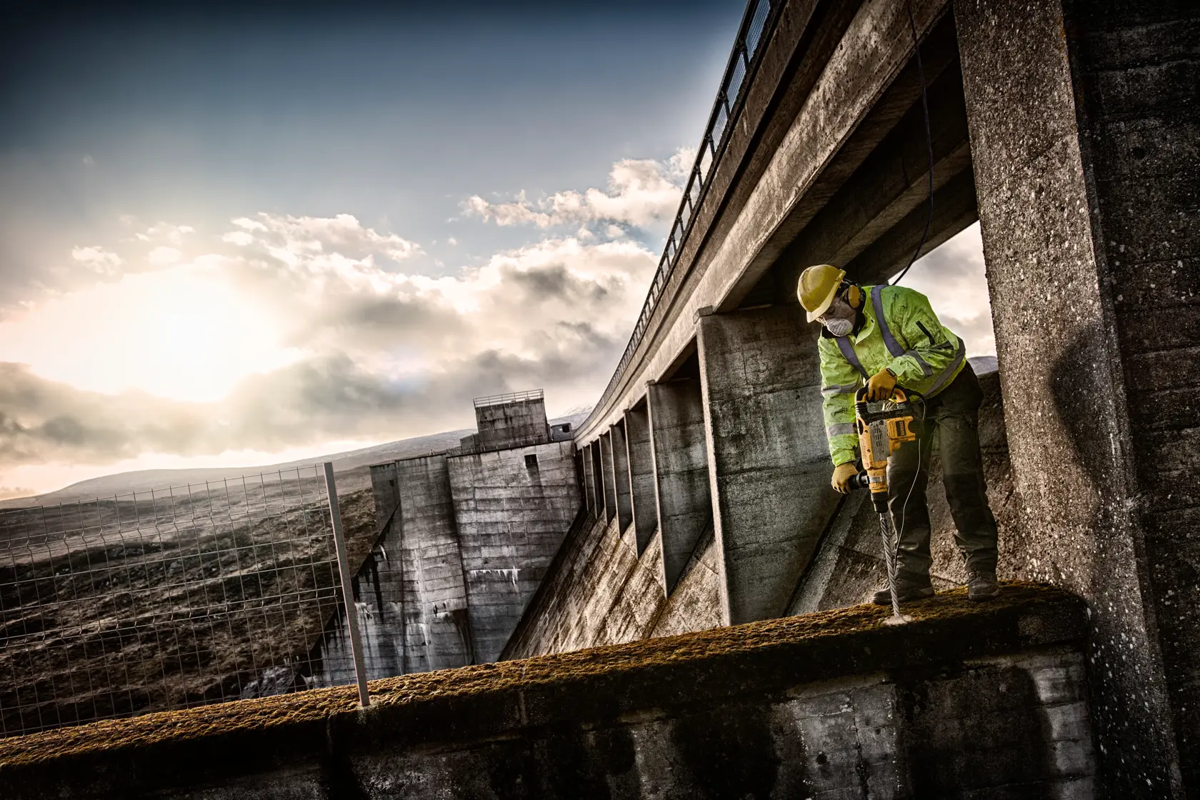 Arbeiter nutzt DEWALT-Bohrer auf Beton bei großer Staumauer und Sonnenuntergang, mit Schutzkleidung.