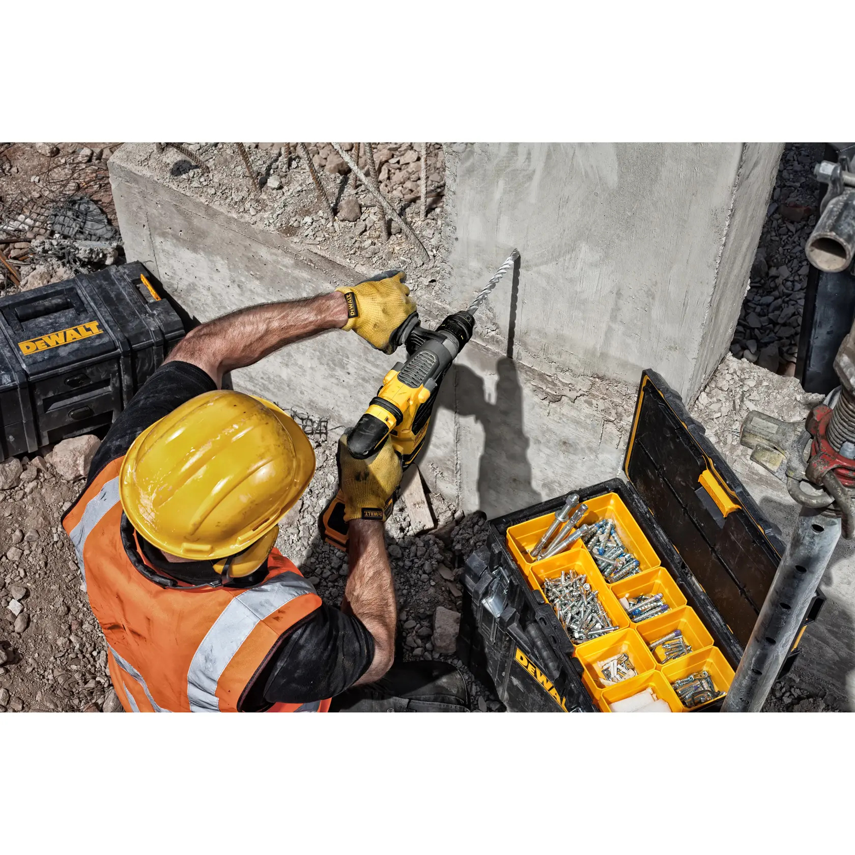 Trabajador perfora pared de concreto con herramienta DEWALT y caja abierta llena de tornillos y pernos.