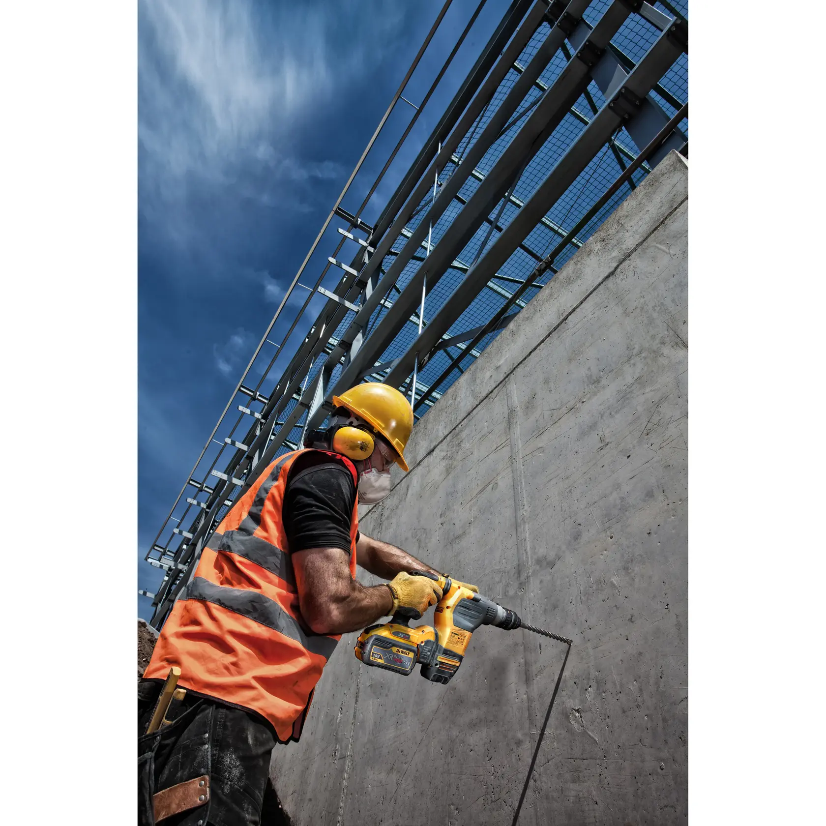 Trabajador con equipo de seguridad usa taladro DEWALT en muro de concreto bajo cielo azul.