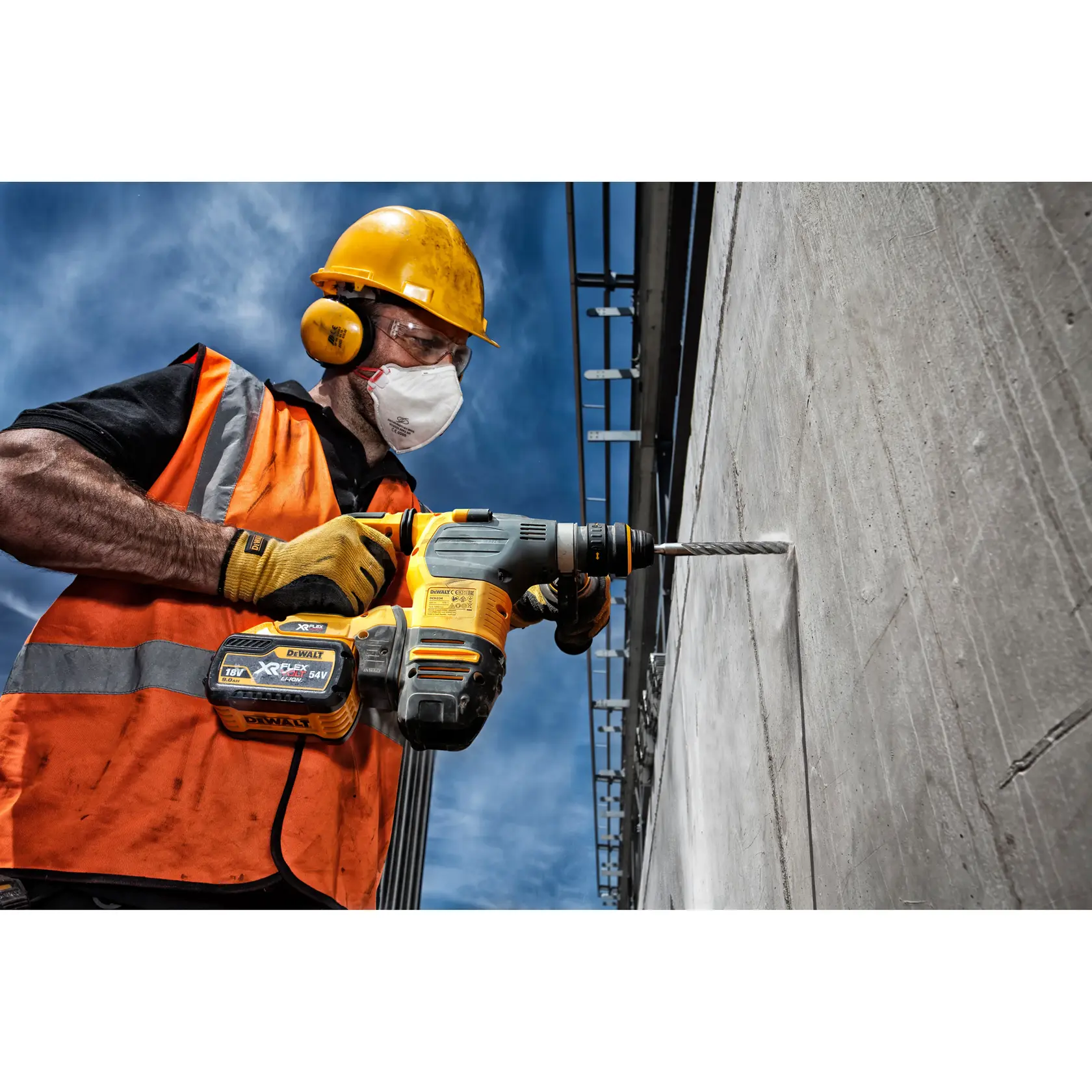Trabajador usa taladro DEWALT en pared de concreto exterior, con equipo de seguridad y chaleco naranja.