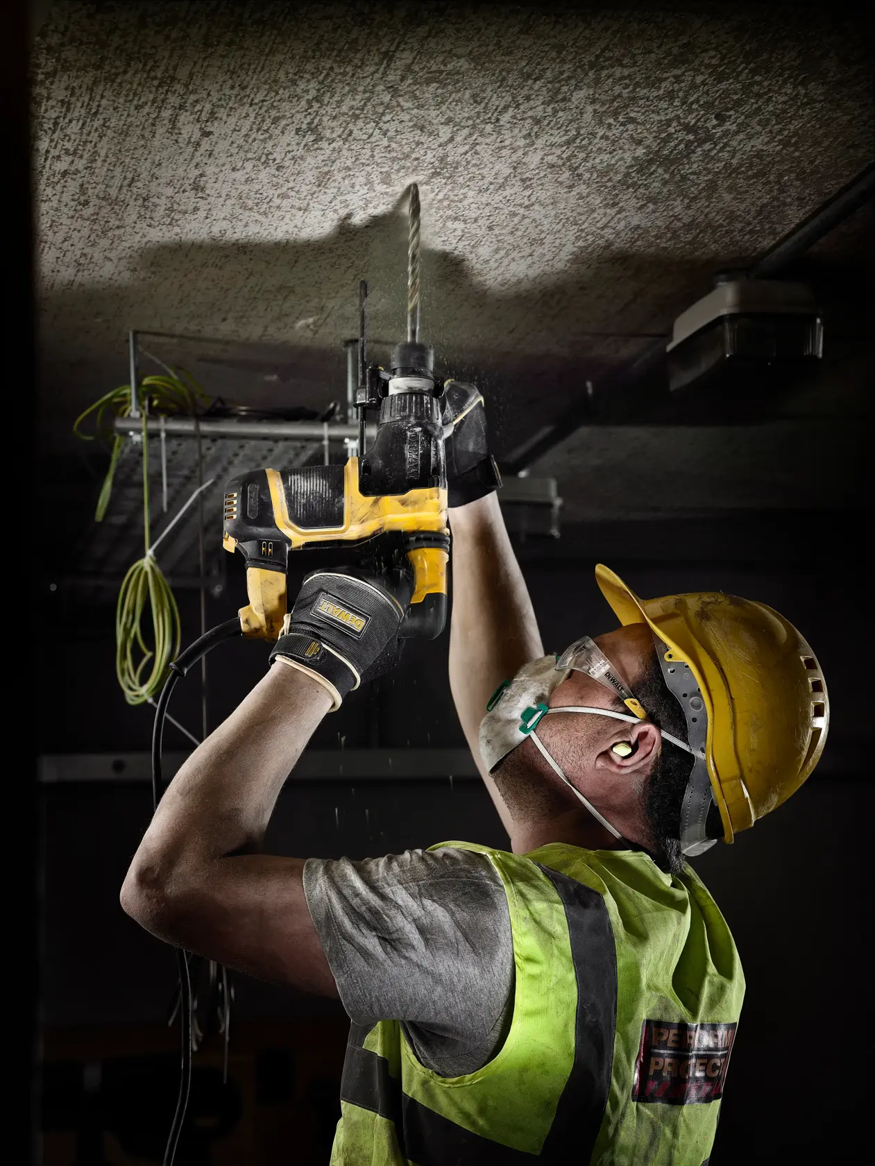 Arbeiter bohrt mit gelbem DEWALT-Werkzeug in die Decke, trägt Sicherheitsausrüstung am Bau.