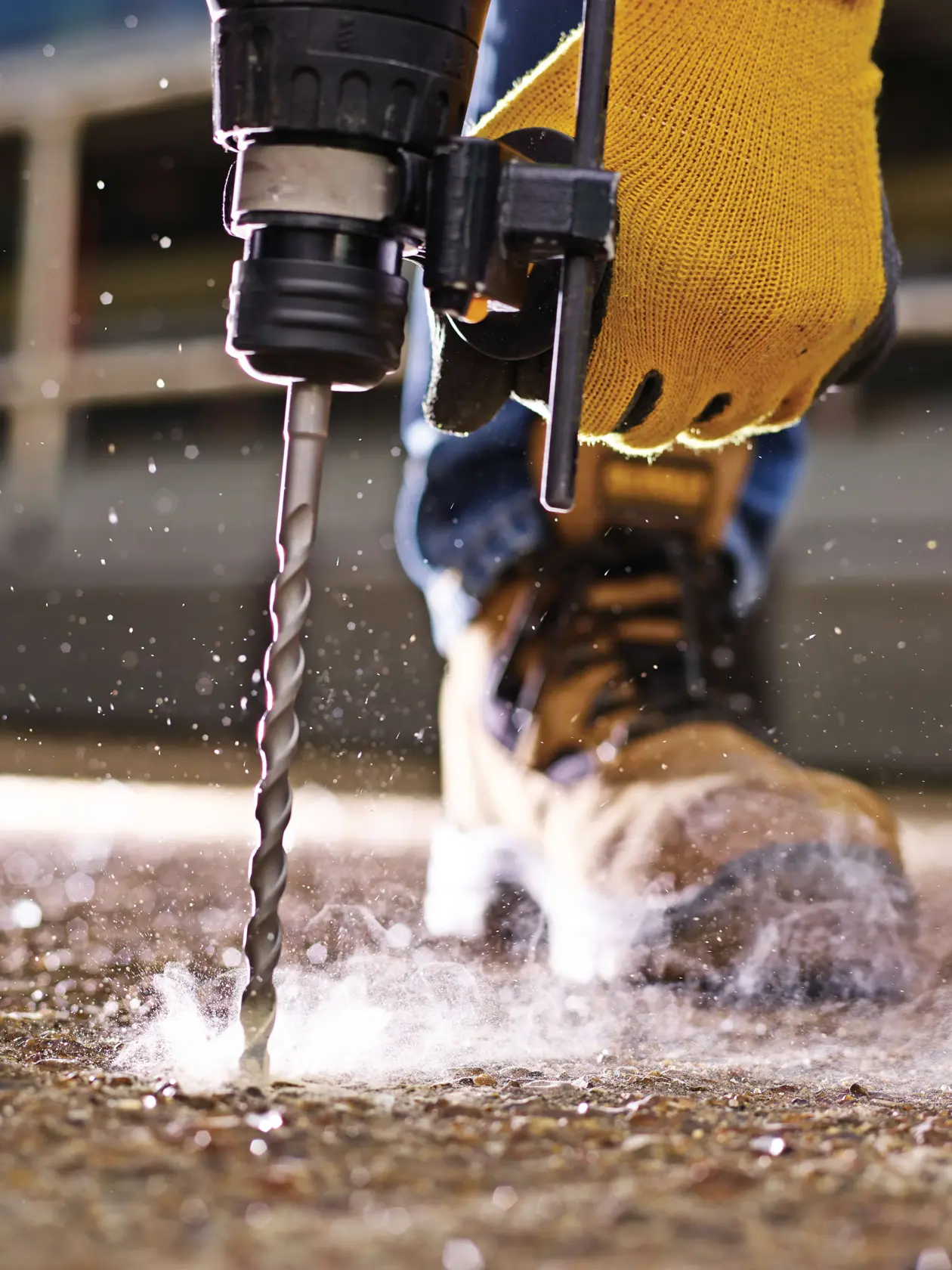Primer plano de mano enguantada usando broca DEWALT en hormigón, polvo y botas de seguridad visibles.