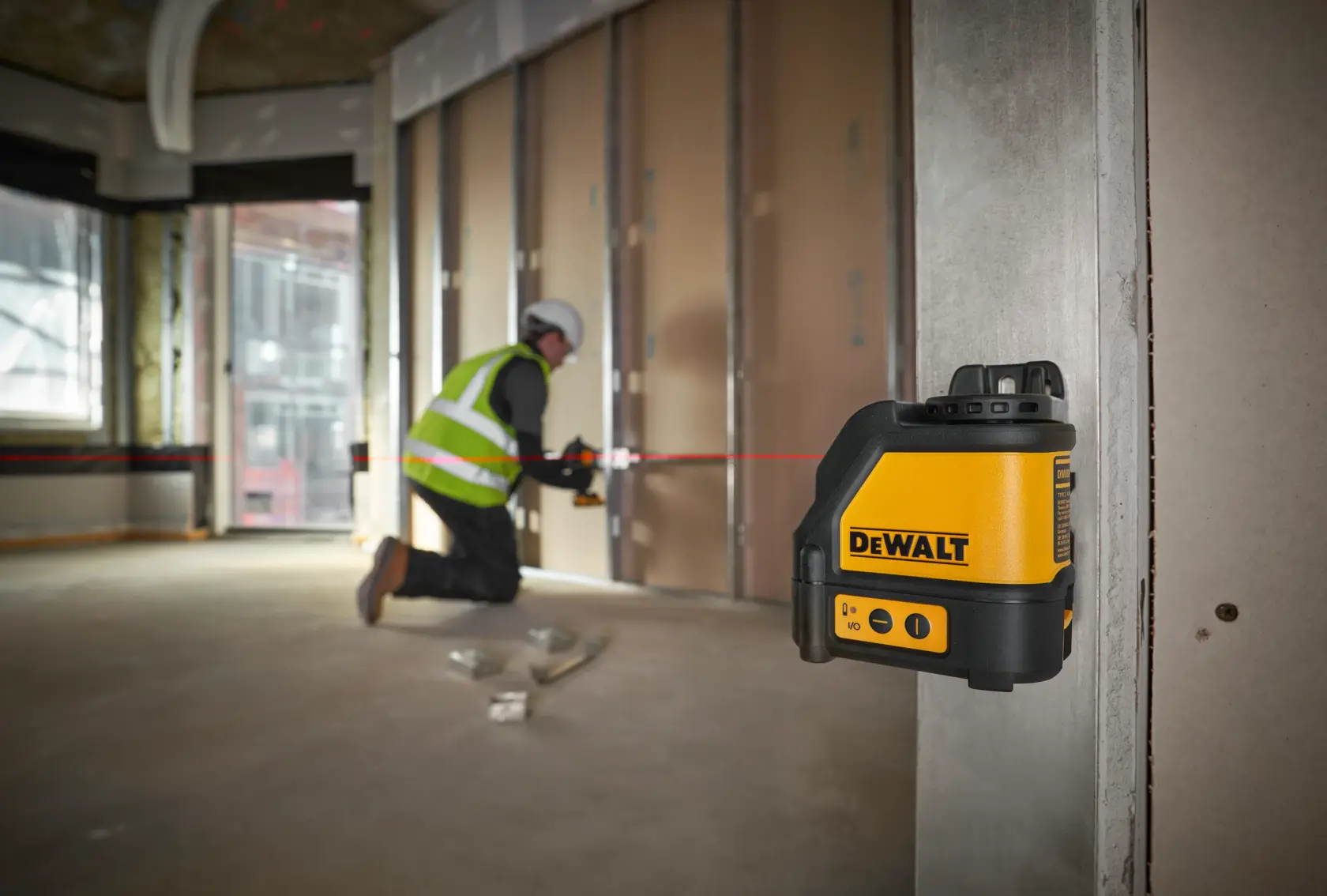DEWALT DW088K Laser-Nivellierer strahlt eine rote Linie im Baugebiet, ein Arbeiter montiert Trockenbauplatten.