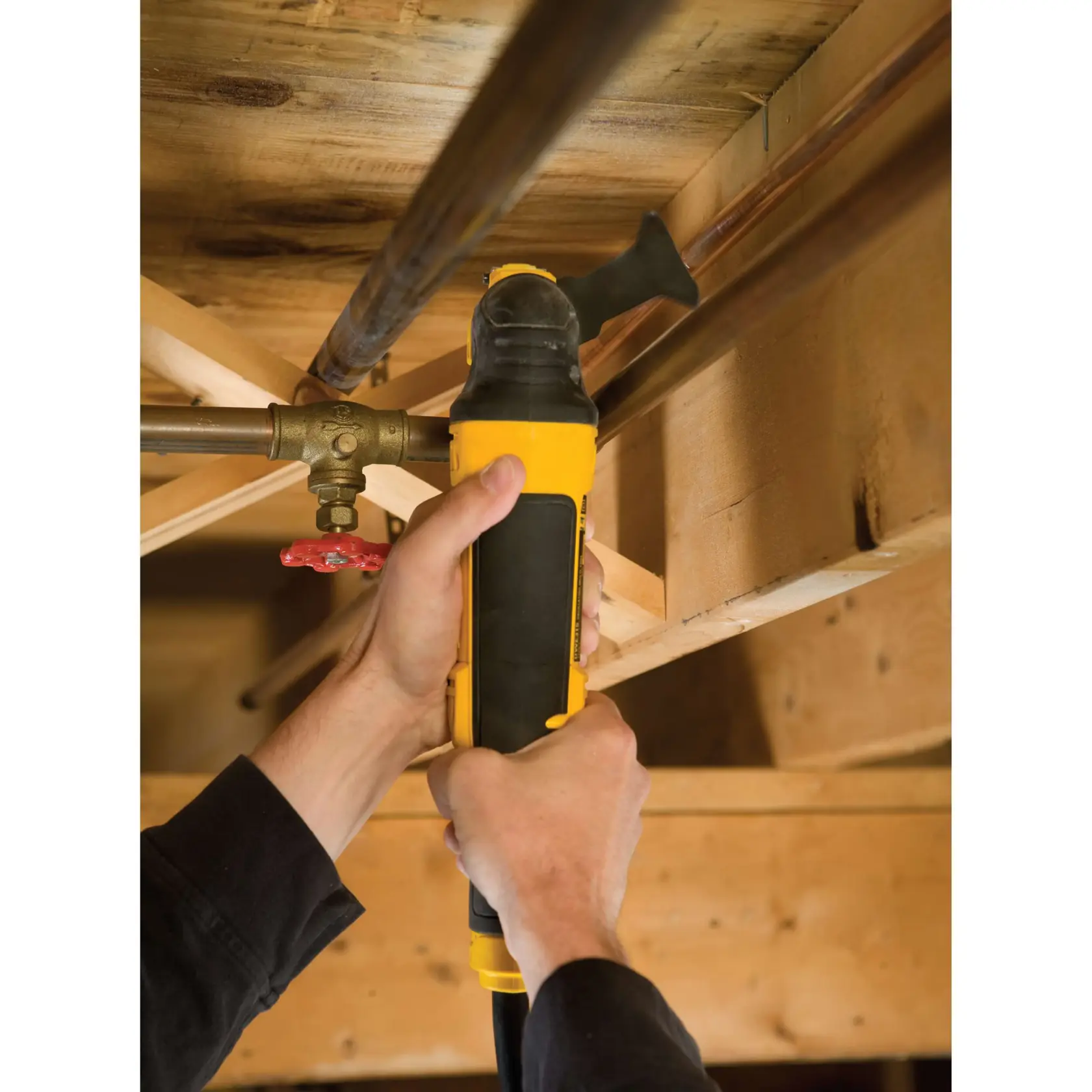 Hände nutzen ein DEWALT Multitool an einem Rohr unter Holzbalken. Gelbes und schwarzes Gerät.