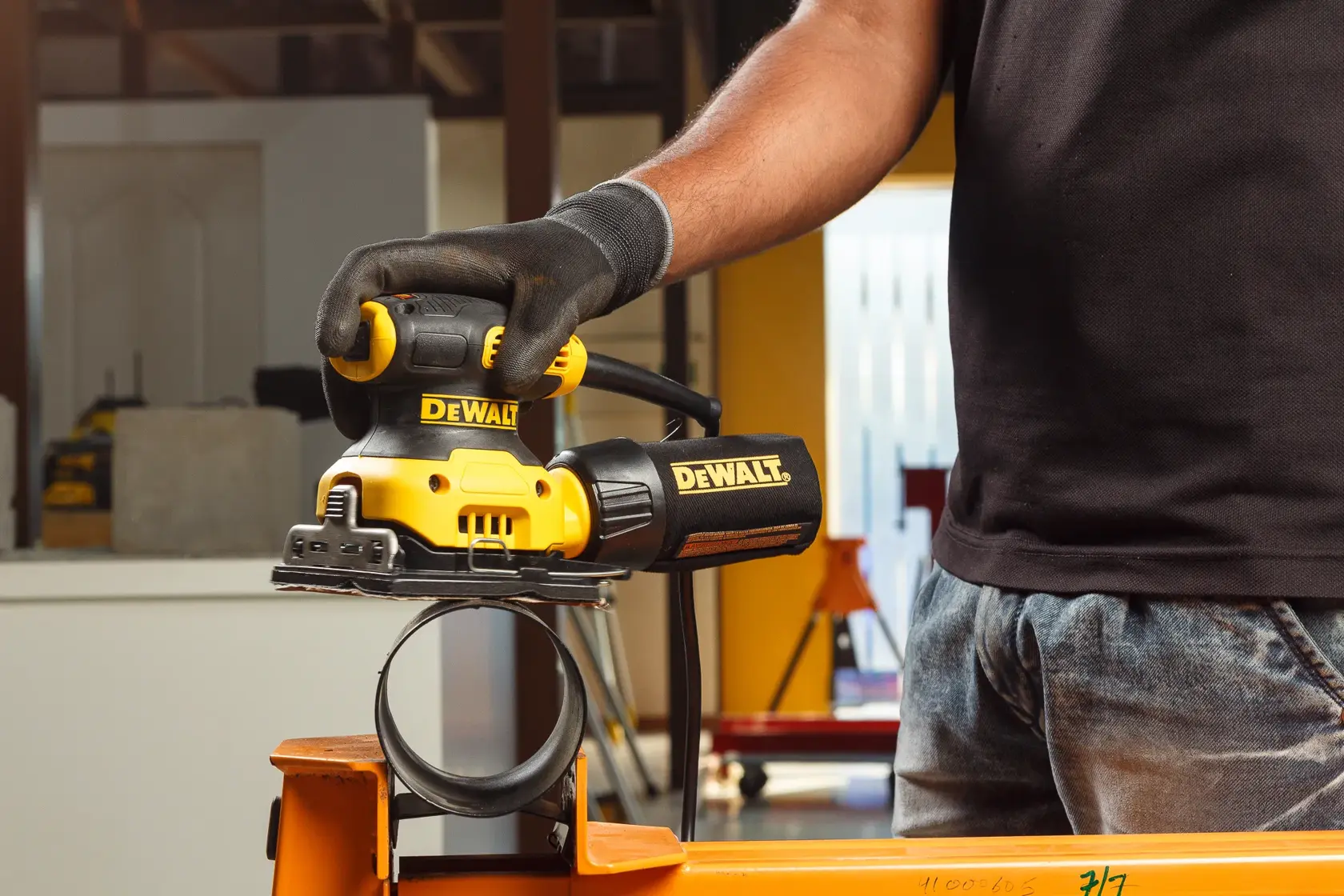 Nahaufnahme einer Person, die einen gelb-schwarzen DEWALT DWE6411 Elektroschleifer verwendet, um eine Holzoberfläche im Freien zu glätten. Die Person trägt Schutzhandschuhe.