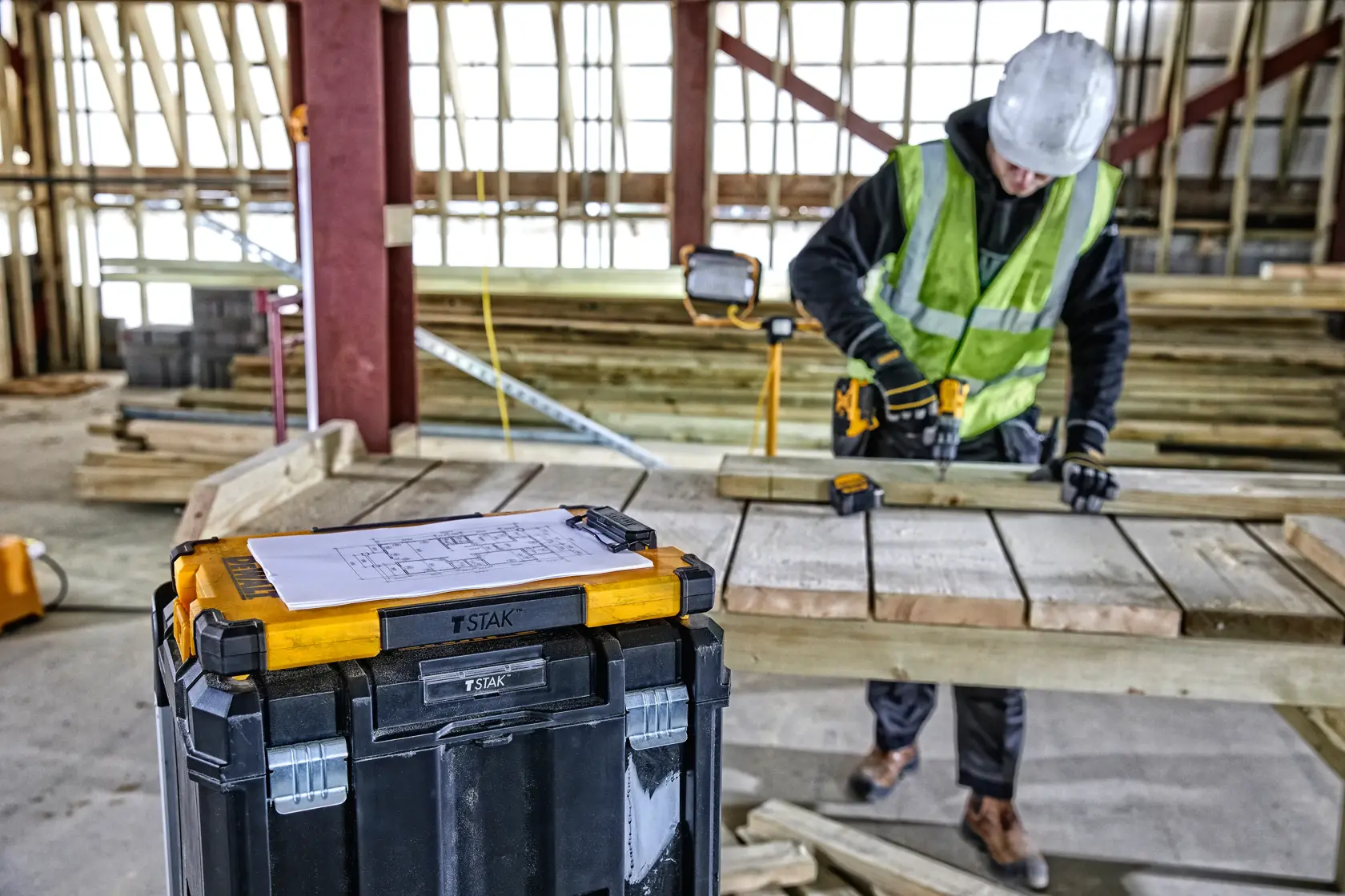 Una caja de herramientas DEWALT TSTAK de diseño amarillo y negro está colocada en primer plano en un sitio de construcción. Encima de la caja hay una hoja de papel con diagramas. Al fondo, un trabajador de la construcción con casco y chaleco de seguridad trabaja con una herramienta eléctrica sobre tablones de madera.