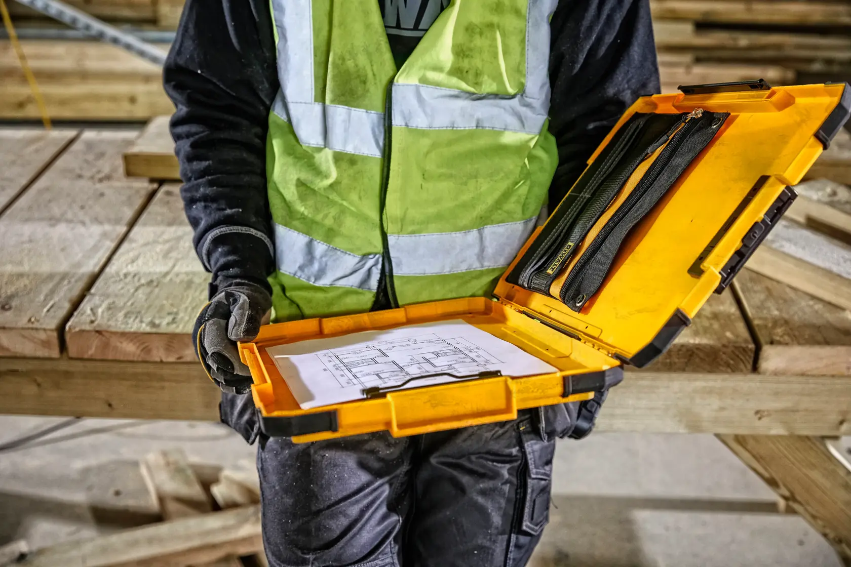 Una persona con un chaleco de seguridad reflectante sostiene un maletín amarillo DEWALT abierto que contiene documentos y herramientas en una obra con vigas de madera al fondo.